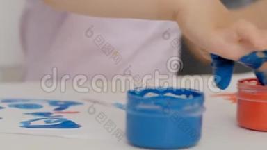 一个小可爱的女孩坐在桌子旁，用明亮的手指颜料画纸，把她的手指浸在颜料罐里。